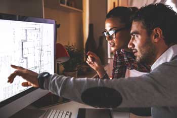 Zwei Personen sitzen vor einem Bildschirm. Eine Person zeigt auf etwas auf dem Bildschirm.